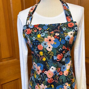 Peut inclure: Tablier floral avec un fond bleu marine fonc&eacute;, orn&eacute; de motifs floraux roses, bleus et rouges avec des feuilles vertes. Le tablier a une encolure am&eacute;ricaine et une poche. Le tablier est port&eacute; sur un t-shirt blanc &agrave; manches longues.