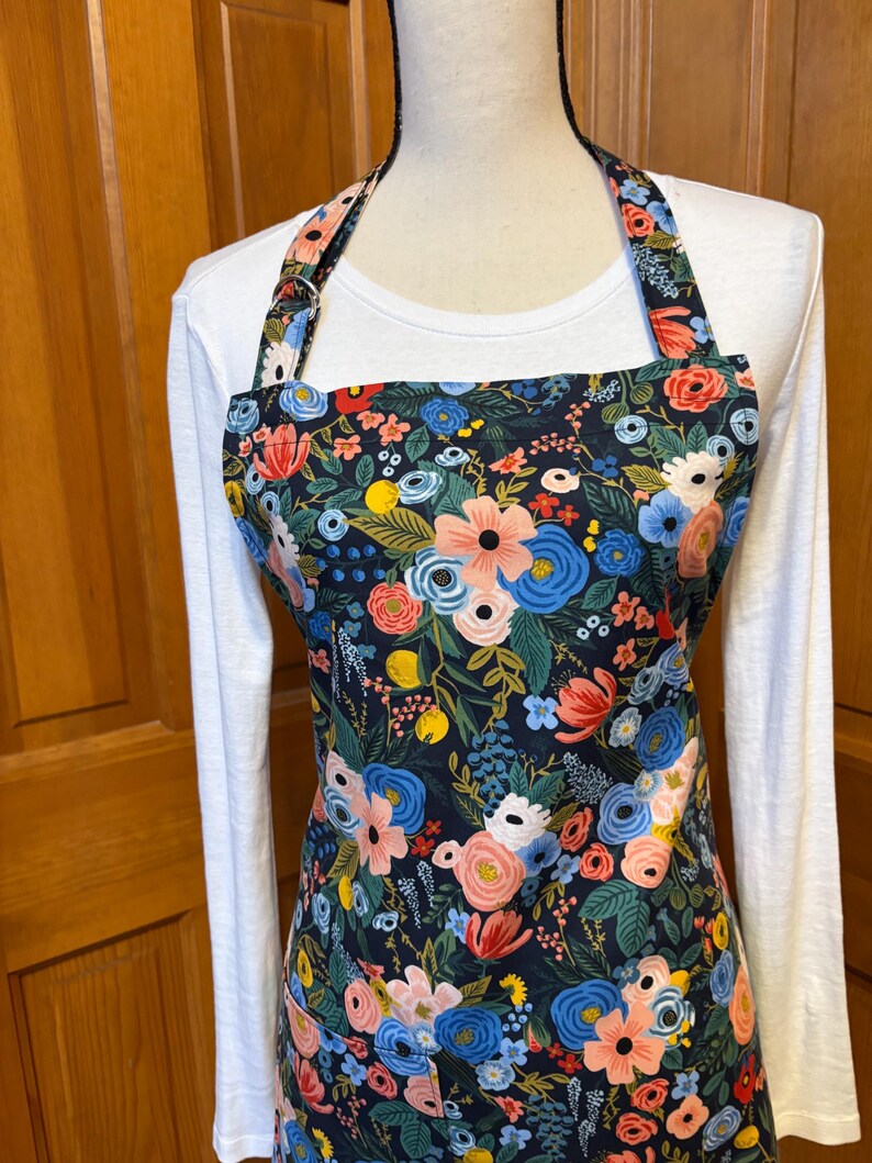Peut inclure: Tablier floral avec un fond bleu marine fonc&eacute;, orn&eacute; de motifs floraux roses, bleus et rouges avec des feuilles vertes. Le tablier a une encolure am&eacute;ricaine et une poche. Le tablier est port&eacute; sur un t-shirt blanc &agrave; manches longues.