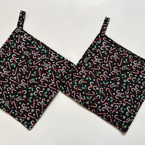 May include: Two square pot holders featuring a black background with a repeating pattern of red and white candy canes, green holly leaves with red berries, and small white snowflakes. Each pot holder includes a small fabric loop for hanging.