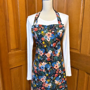 Peut inclure: Tablier floral avec un fond bleu marine fonc&eacute;, orn&eacute; de fleurs roses, bleues et jaunes. Le tablier est pr&eacute;sent&eacute; sur un mannequin, mettant en valeur son design. Il poss&egrave;de un col halter et une poche.