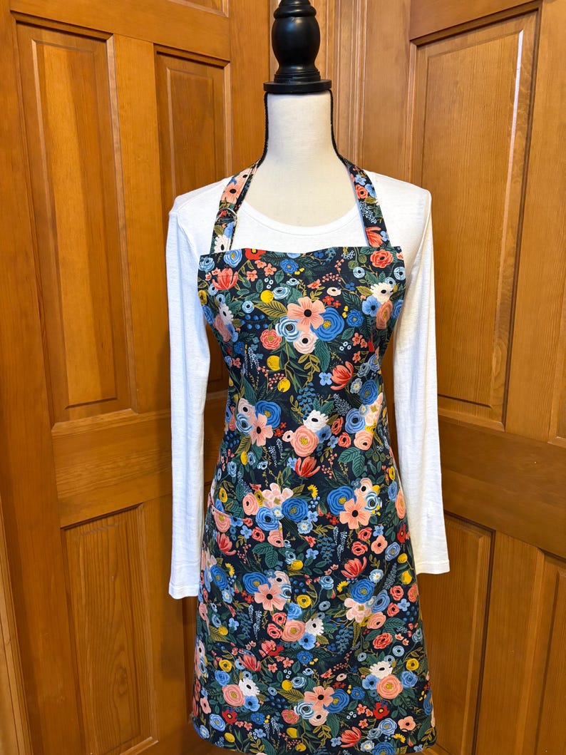 Peut inclure: Tablier floral avec un fond bleu marine fonc&eacute;, orn&eacute; de fleurs roses, bleues et jaunes. Le tablier est pr&eacute;sent&eacute; sur un mannequin, mettant en valeur son design. Il poss&egrave;de un col halter et une poche.