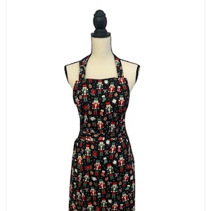 May include: A black apron with a festive Christmas pattern. The apron features a repeating design of nutcrackers, gingerbread men, and other holiday-themed elements in red, green, and white. The apron has a halter neck and a waist tie.