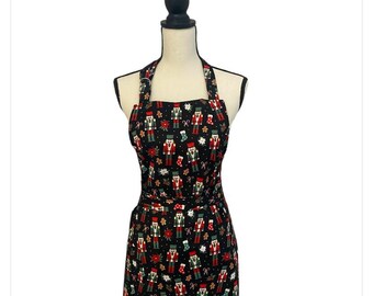 May include: A black apron with a festive Christmas pattern. The apron features a repeating design of nutcrackers, gingerbread men, and other holiday-themed elements in red, green, and white. The apron has a halter neck and a waist tie.