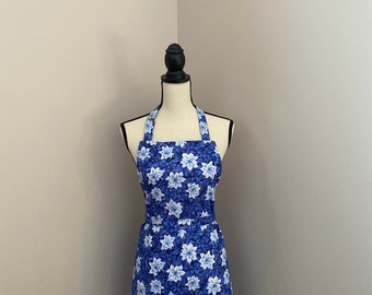 May include: A blue and white floral print apron displayed on a mannequin. The apron features a halter neck, a fitted bodice, and a skirt that falls to the knees. The floral pattern includes white flowers on a blue background.