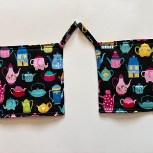 May include: Two square quilted pot holders featuring a black background with a colorful repeating pattern of various teapots, kettles, and small houses in shades of pink, blue, yellow, and green. Each pot holder includes a fabric loop for hanging.
