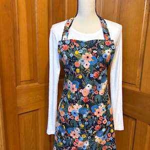 Peut inclure: Un tablier floral avec un fond bleu marine fonc&eacute;, pr&eacute;sentant un motif de fleurs roses, bleues et jaunes. Le tablier a un col licou et une forme longue et rectangulaire, expos&eacute; sur un mannequin.