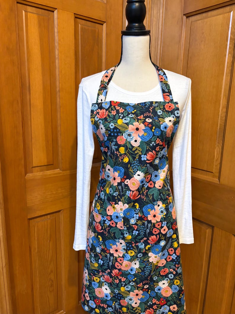 Peut inclure: Un tablier floral avec un fond bleu marine fonc&eacute;, pr&eacute;sentant un motif de fleurs roses, bleues et jaunes. Le tablier a un col licou et une forme longue et rectangulaire, expos&eacute; sur un mannequin.