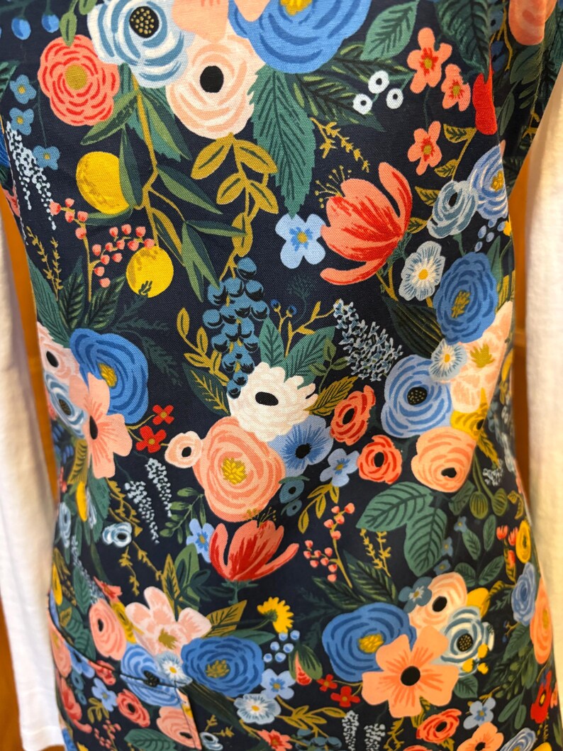 Peut inclure: Un tablier bleu marine orn&eacute; d'un motif floral vibrant. Le design pr&eacute;sente un assortiment de fleurs color&eacute;es dans des tons de rose, de bleu et de corail, avec des feuilles vertes et des fruits jaunes. Le tablier est en tissu r&eacute;sistant.