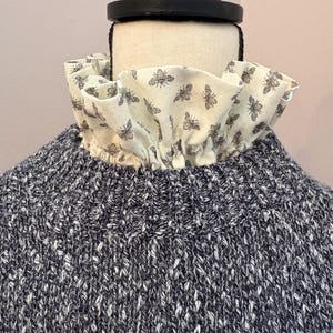 May include: A close-up of a blue and white knit sweater with a cream-colored ruffled collar. The collar is patterned with small, gray bee illustrations. The sweater is displayed on a mannequin.
