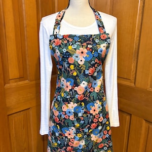 Peut inclure: Tablier floral avec un fond sombre, pr&eacute;sentant un motif de fleurs roses, bleues et jaunes. Le tablier a un bavoir, des liens &agrave; la taille et une sangle de cou. Le tablier est pr&eacute;sent&eacute; sur un mannequin portant un t-shirt blanc &agrave; manches longues.