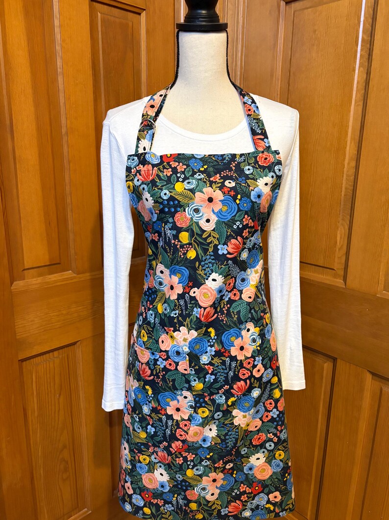 Peut inclure: Tablier floral avec un fond sombre, pr&eacute;sentant un motif de fleurs roses, bleues et jaunes. Le tablier a un bavoir, des liens &agrave; la taille et une sangle de cou. Le tablier est pr&eacute;sent&eacute; sur un mannequin portant un t-shirt blanc &agrave; manches longues.