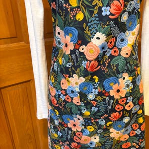 Peut inclure: Un tablier &agrave; imprim&eacute; floral sur fond bleu marine fonc&eacute;, orn&eacute; de fleurs roses, bleues, jaunes et blanches. Le tablier est dot&eacute; d'un bavoir et de poches. Il est port&eacute; sur un haut blanc &agrave; manches longues.