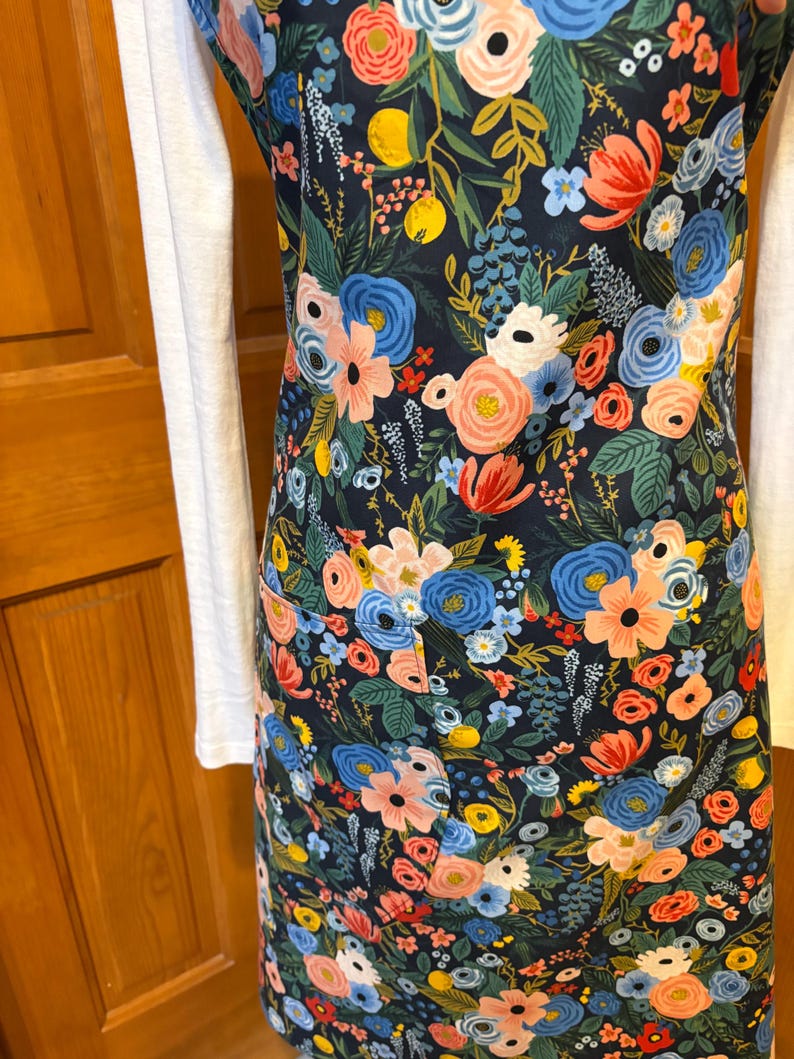 Peut inclure: Un tablier &agrave; imprim&eacute; floral sur fond bleu marine fonc&eacute;, orn&eacute; de fleurs roses, bleues, jaunes et blanches. Le tablier est dot&eacute; d'un bavoir et de poches. Il est port&eacute; sur un haut blanc &agrave; manches longues.