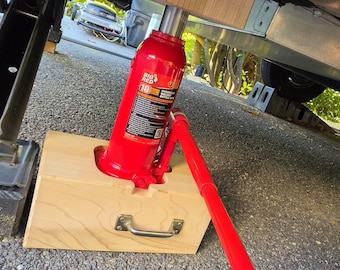 May include: A red Big Red 10-ton hydraulic bottle jack positioned on a light-colored wooden base with a metal handle. The jack extends upward to a wooden block labeled JACK, supporting the underside of a vehicle frame with a long red handle.