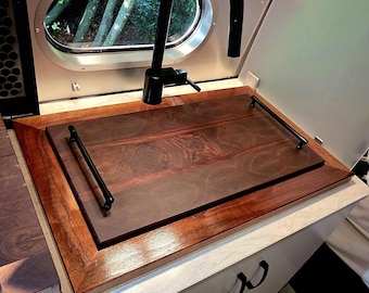 May include: Interior view of a modern RV kitchen with a dark wood countertop and a black faucet. The countertop has a built-in sink and a wooden cover. The windows offer a view of the outside. The cabinet has black handles.
