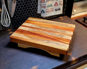 May include: A rectangular wooden cutting board with a light and dark wood grain pattern. The board is elevated on a small base. The wood tones include light tan, medium brown, and reddish-brown stripes. Kitchen and dining decor.