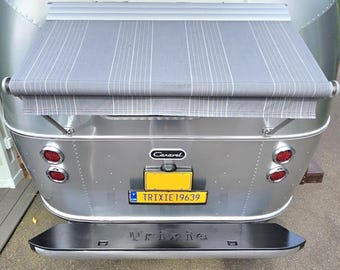 May include: The rear view of a silver Airstream Caravel travel trailer featuring a grey striped window awning, a yellow license plate reading TRIXIE19639, and a black rear bumper with the name Trixie embossed in the center of the metal surface.
