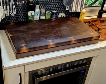 May include: A dark wood butcher block countertop sits above a microwave oven in a kitchen setting. The countertop is rectangular and appears to be made of end-grain wood. Utensils hang above the countertop, and bottles of olive oil and spices are visible.