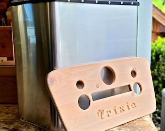 May include: A stainless steel cooler with a black lid and a light-colored wooden lid with the name "Trixie" cut out. The wooden lid has several circular cutouts. The cooler is sitting on a wooden surface.
