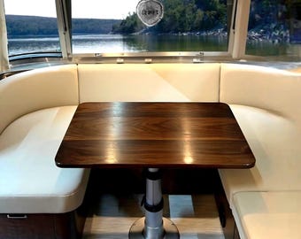 May include: A dark brown wooden table with a smooth, polished surface and a central silver metal pedestal base, positioned between two cream-colored upholstered bench seats inside a vehicle with a large window view of a lake.