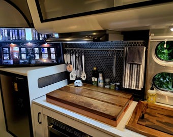 May include: A small kitchen in a camper van with a wood cutting board, a stainless steel range hood, and a black and white tiled backsplash. There are various kitchen utensils hanging on a rack above the cutting board.