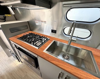 May include: Interior view of a modern RV kitchen. Features include a stainless steel sink, a gas stovetop, and a microwave oven. The countertop is made of wood. The walls are stainless steel, and there are oval windows.