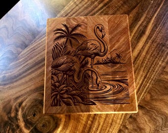 May include: A square wooden cutting board with a laser-etched design of two flamingos in a tropical setting. The flamingos are near water with palm trees in the background. The wood has a rich, warm tone.