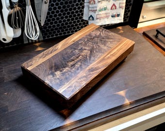 May include: A rectangular wooden cutting board with a dark brown center and lighter brown edges. The wood grain is visible. The board is on a dark surface, with kitchen utensils hanging in the background. A second cutting board is also visible.