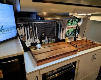 May include: A small kitchen with white cabinets and a stainless steel sink. There are two wooden cutting boards on the counter, one with a copper edge. A television is mounted on the wall above the counter. There are various kitchen utensils hanging on a rack above the counter.