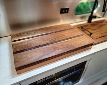 May include: A large, rectangular wooden cutting board with a light brown finish and two dark brown walnut strips running lengthwise. The board is made of wood with a natural grain pattern.