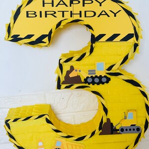 May include: Yellow construction themed number 3 pinata with black and yellow striped paper. The pinata has the words "Happy Birthday" on the top and features construction vehicles.