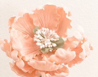 May include: A single peach-colored artificial flower with a cluster of white pearl-like accents in the center. The flower has a green stem and is in a clear glass vase.