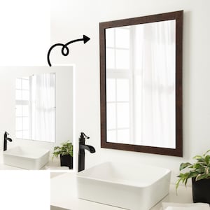 May include: A rectangular mirror with a dark brown wooden frame is mounted on a white wall. The mirror reflects a window with white curtains. A white rectangular sink and black faucet are below the mirror.