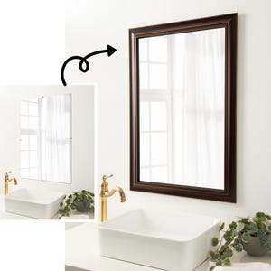 May include: A framed bathroom mirror with a dark brown frame. The mirror is reflecting a white bathroom with a sink and faucet.