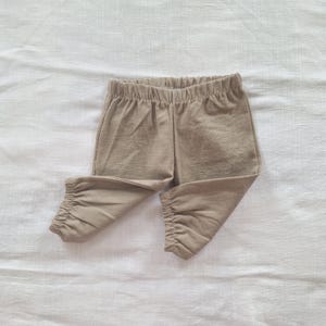 May include: A pair of solid tan pants featuring an elasticized waistband and gathered elastic cuffs at the bottom of each leg. The pants are crafted from a textured fabric with a visible weave, providing a simple and functional design for daily wear.