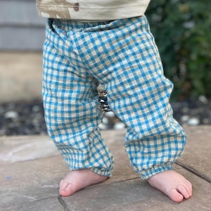 May include: Blue and beige gingham baby pants with elastic cuffs and waistband. The pants are part of a baby outfit. The baby is standing on a stone surface.