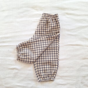 May include: A pair of baby pants in a neutral color palette. The pants feature a classic gingham pattern in shades of beige and gray. The waist and ankles are elasticized for a comfortable fit. The fabric appears soft and lightweight.