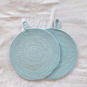 May include: Two round, light blue coasters or trivets crafted from a thick, coiled rope material. Each circular piece features a spiral pattern radiating from the centre and includes a small, matching rope loop at the top for convenient hanging.