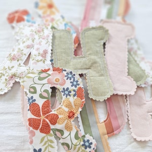 May include: A collection of fabric letters featuring various patterns and solid colors. The letters include floral prints in red, pink, and blue, solid sage green, soft pink, and striped designs in muted earth tones, all with scalloped stitched edges.