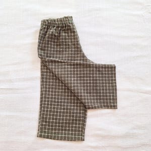 May include: Olive green pants featuring a white grid pattern throughout. The pants have a straight-leg cut and an elasticized waistband at the top. The fabric displays a visible woven texture, and the garment is presented in a flat, folded position.
