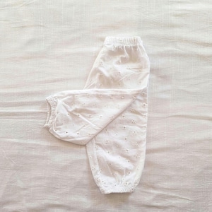 May include: A pair of white pants featuring an all-over eyelet embroidery pattern. The garment has an elasticized waistband and elastic cuffs at the bottom of each leg, creating a gathered, tapered silhouette with a lightweight appearance.