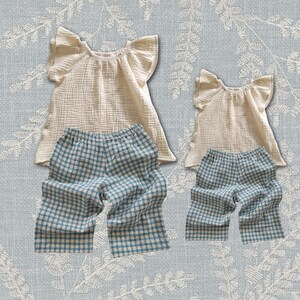 May include: Two sets of clothing, each consisting of a cream-coloured, textured, short-sleeved top with ruffled shoulders and a pair of blue and white gingham-patterned trousers with an elastic waistband, displayed in two different sizes.