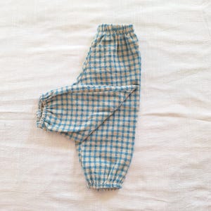 May include: A pair of pants featuring a blue and beige gingham check pattern. The garment has an elasticized waistband and elasticized cuffs at the ankles, creating a gathered, tapered silhouette with a consistent checkered design throughout.