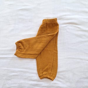 May include: Mustard yellow pants made of a textured, crinkled fabric. The pants feature an elasticized waistband and elasticized cuffs at the bottom of each leg, creating a gathered, tapered silhouette with a soft, lightweight appearance.