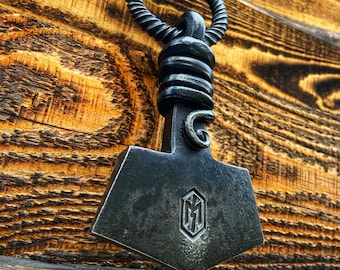 May include: A black metal necklace with a hammered,  M-shaped pendant. The pendant is shaped like a small axe head. The necklace is hanging on a wooden surface.