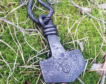 May include: A black metal hammer with a twisted handle and a decorative design on the head. The hammer is lying on a bed of green moss.