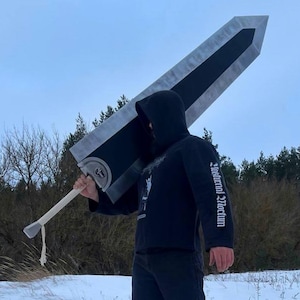 May include: A large, black and silver sword prop with a long, light-colored handle is held by a person in a black hooded sweatshirt and black pants. The person is standing in a snowy field with trees in the background. The sword has a distinctive, angular blade.