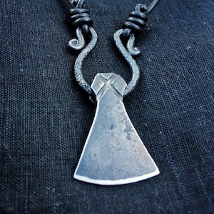 May include: A silver axe pendant necklace with a black leather cord. The axe head has a cross-hatched design and a smooth, slightly curved blade.