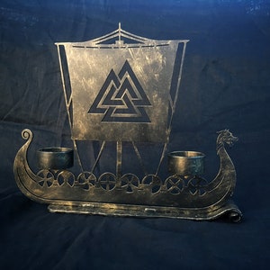 Könnte beinhalten: Ein Kerzenhalter aus Metall in Form eines Wikingerschiffs mit einem Valknot-Design auf dem Segel. Das Schiff hat drei Kerzenhalter und eine goldene Oberfläche.