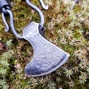 May include: A black metal axe pendant with a leather cord. The pendant is on a bed of green moss.